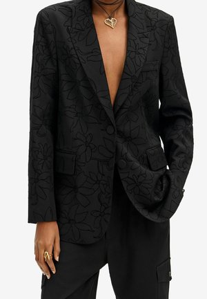 Black blazer with subtle floral pattern worn open over chest, paired with black pants, gold leaf necklace, and chunky gold ring on left hand.