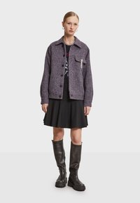 Lilla hørjacket med krave og frontlomme, parret med en sort nederdel og høje sorte platformstøvler. Sølvkors detalje på lommen.