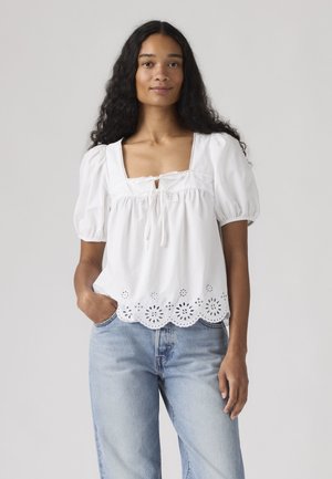 Jonge vrouw met lang krullend haar draagt een wit blouse met pofmouwen en geborduurde zoom, gecombineerd met lichtblauwe jeans, staand tegen een effen achtergrond.