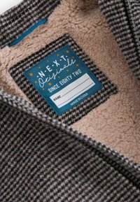Étiquette en tissu bleu sur une veste à carreaux noir et blanc doublée en polaire, indiquant « NEXT Originals since eighty two », avec un champ de nom vierge.