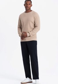 Beige strukturierter Strickpullover mit Rundhalsausschnitt, kombiniert mit dunkelblauen Hosen und weißen Sneakers mit braunen Akzenten.