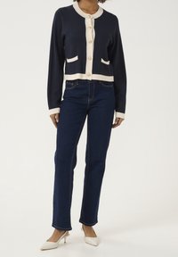 Marineblauer Cardigan mit cremefarbenen Akzenten, runder Ausschnitt, Knopfleiste und zwei vorderen Taschen, kombiniert mit blauen Jeans und weißen High Heels.