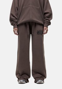 Brauner, oversized Kapuzenpullover mit Reißverschluss und passenden, weit geschnittenen Sweatpants, die ein schwarzes Grafikdesign auf dem linken Oberschenkel zeigen. Weiches Stoffgefühl.