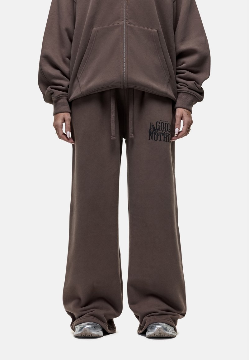 Brauner, oversized Kapuzenpullover mit Reißverschluss und passenden, weit geschnittenen Sweatpants, die ein schwarzes Grafikdesign auf dem linken Oberschenkel zeigen. Weiches Stoffgefühl.