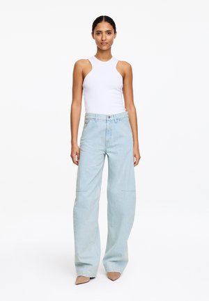 Kobieta w białym dopasowanym topie bez rękawów, jasnoniebieskich szerokich dżinsach i beżowych spiczastych butach, stojąca na tle jednolitej białej ściany.