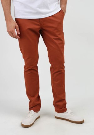 Chinos couleur rouille en coton, avec une coupe slim, des poches latérales et un design à l'avant plat, associés à des chaussures de sport blanches.
