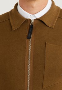 Hnědá pletená košile s texturou a zapínáním na zip a kapsou na prsou. Obsahuje bílou košili s límečkem, která kontrastuje s hnědou.
