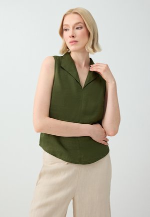 Femme blonde aux cheveux courts portant un haut sans manches vert foncé à col et un pantalon beige clair, regardant vers sa gauche les bras croisés.