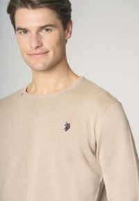 Maglione beige a maglia con scollatura rotonda, caratterizzato da un piccolo emblema blu navy sul petto e una sottile trama a costine lungo i polsini.