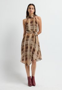 Vrouw in een mouwloze, hoge hals, bruin en beige patroon jurk met een asymmetrische zoom en een geknoopte taille, gecombineerd met rode enkellaarsjes.