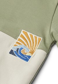Camicia a manica corta in tessuto di cotone verde con orlo crema, caratterizzata da una toppa ricamata di onde e sole nei colori blu, giallo e bianco.