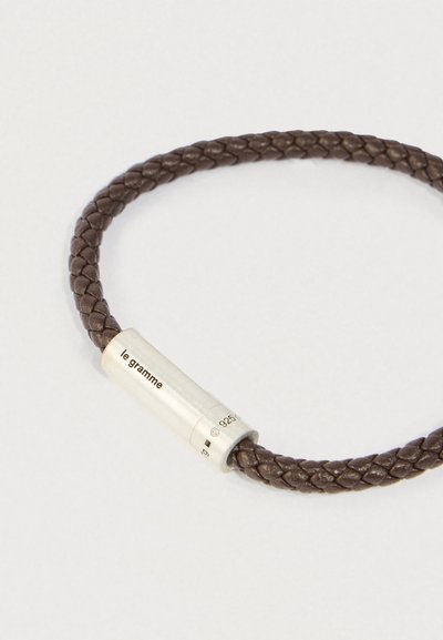 Brūns pītas ādas aproce ar sudraba cilindrisku sprādzi, uz kuras ir iegravēts "le gramme" un "925" zīmogs.