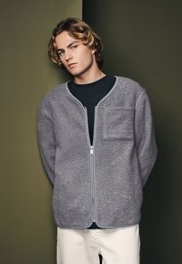 Jachetă din fleece gri cu guler rotund, fermoar frontal și un buzunar pe piept. Are un material texturat și detalii de contrast.