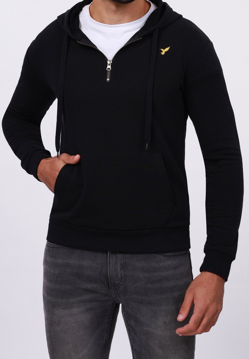 Homme portant un sweat à capuche noir zippé avec cordons de capuche, logo d'oiseau jaune, t-shirt blanc et jean gris foncé, main dans la poche du sweat.