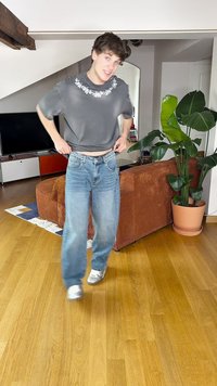 Camisa gris de manga corta con bordado floral, jeans holgados de un azul claro y zapatillas blancas, de pie sobre un suelo de madera con plantas y muebles de interior.