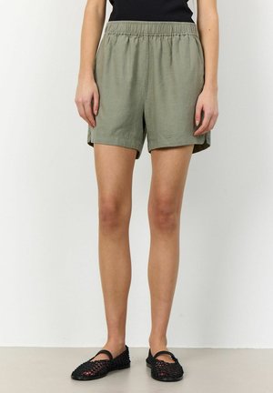 Shorts en lin vert clair avec une taille élastique et des poches latérales, présentant une coupe décontractée et des ourlets roulés. Associés à des chaussures noires à enfiler.