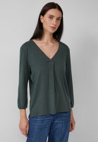 Femme avec des cheveux noirs lisses portant un haut à manches longues en V vert foncé et un jean bleu, posant contre un fond gris clair uni.