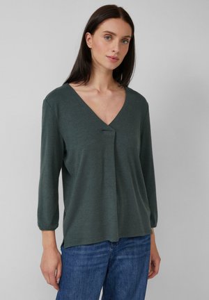 Eine Frau mit glattem, dunklem Haar, die ein dunkelgrünes V-Ausschnitt-Langarmoberteil und blaue Jeans trägt, posiert vor einem einfarbigen hellgrauen Hintergrund.