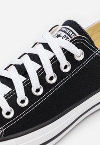 Baskets Converse basses noires avec des lacets blancs, œillets en métal et semelle en caoutchouc blanc présentant des rayures noires doubles.
