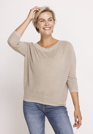 Lächelnde Frau mit kurzen blonden Haaren, die einen beigen lockeren Pullover und blaue Jeans trägt, und mit einer Hand ihr Haar berührt.