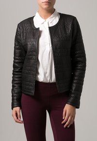 Svart quiltad jacka med geometriskt mönster, rund krage och åtsittande ärmar, i kombination med en vit skjorta och mörka leggings.