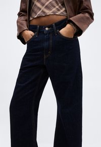 Personne portant un jean large taille haute bleu foncé avec les mains dans les poches, un crop top à carreaux et une veste en cuir marron.