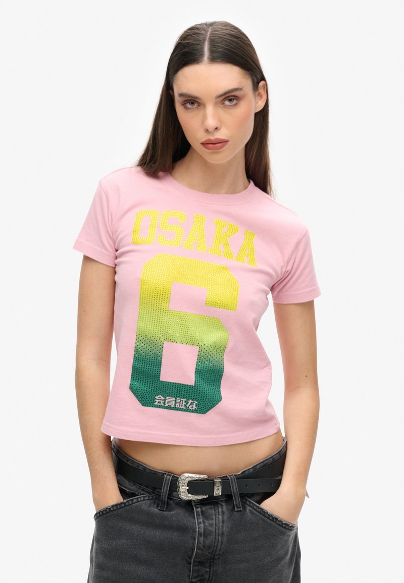 Rosa Cropped T-Shirt mit gelbem und grünem Farbverlauf "OSAKA 6" Druck und strukturierten Details. Kombiniert mit dunklen Jeans und einem schwarzen Gürtel.