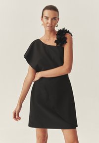 Fekete ruha aszimmetrikus, egy válaszos dizájnnal, fodros virágos díszítéssel, sima textúrával és térd alá érő szilhuettel.