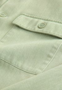 Gros plan sur un tissu vert clair avec une poche à rabat boutonnée et des coutures visibles sur le matériau texturé.