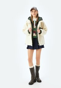 Fluffy crèmekleurige jas over een groene rits-hoodie, gestreepte blouse, marineblauwe rok en bruin kniehoge laarzen met gespen. Een grijze pet maakt de look compleet.