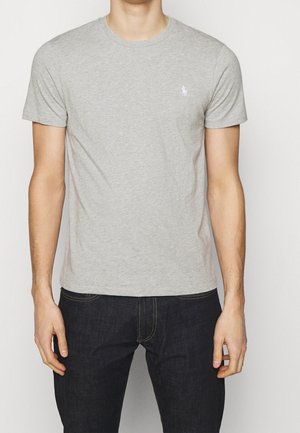 Hombre que lleva una camiseta lisa de manga corta de color gris claro con un pequeño logo blanco bordado en el pecho izquierdo y vaqueros oscuros.