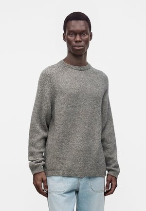 Homme portant un pull en maille gris et un jean bleu clair, debout devant un fond blanc uni.