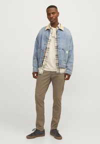 Uomo in piedi che indossa una giacca di jeans azzurro chiaro, una camicia a quadri beige, una maglietta color crema, pantaloni marroni e scarpe nere con suole in gomma.