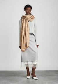 Μπεζ κασκόλ με διακριτό σχέδιο, ριγμένο πάνω σε ένα ανοιχτό γκρι πλεχτό πουλόβερ και σε μία μακριά γκρι φούστα με λευκή δαντελένια ταινία. Οι λευκές γόβες ολοκληρώνουν το ντύσιμο.