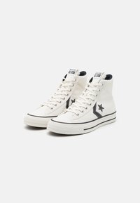 Adidași cu prindere înaltă din pânză de culoare albă murdară, cu accente negre, având un logo în formă de stea, șireturi tradiționale și talpă din cauciuc texturat.
