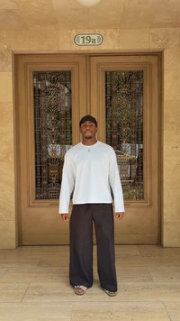 Weißes Langarmhemd, lockere schwarze Hose und Sandalen; steht vor kunstvoll verzierten Doppeltüren mit floraler Metallarbeit und einer Nummerntafel.