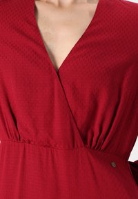 Robe rouge à encolure en V profonde, taille froncée, tissu texturé avec des motifs subtils et un petit détail métallique.
