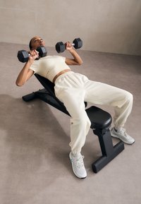 En person ligger på en svart vektbenk og løfter heksagonale manualer. De har på seg en kremfarget crop top og matchende joggebukser, med grå joggesko.
