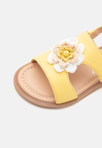 Sándalo sintético amarillo con adornos florales en crema y amarillo. Presenta puntera abierta, correa ajustable y suela plana.