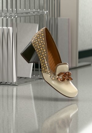 Zapato de tacón cuadrado en crema y con estampado, decorado con una cadena de carey, colocado sobre un suelo reflectante cerca de una estantería de metal y paneles blancos.