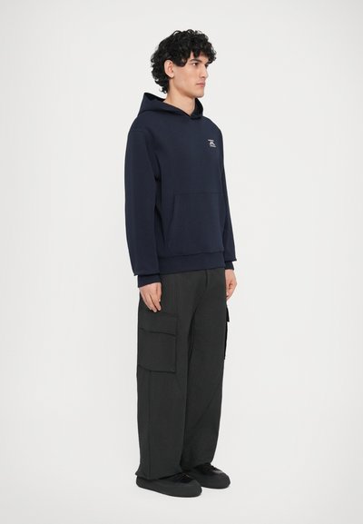 Μπλε σκουρό φούτερ με κουκούλα και μπροστινή τσέπη, διακοσμημένο με διακριτικό λογότυπο. Συνδυασμένο με άνετο, μαύρο cargo παντελόνι με πλαϊνές τσέπες και μαύρα παπούτσια.