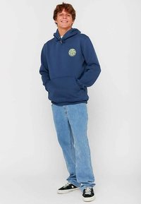 Felpa con cappuccio blu navy con tasca frontale, toppa con logo, abbinata a jeans azzurri e sneakers nere con suole bianche.