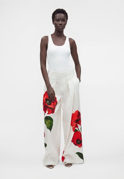 Femme portant un haut blanc sans manches et un pantalon blanc à jambes larges avec de grandes impressions florales rouges, debout avec une main dans la poche.