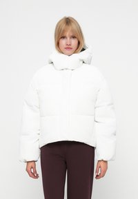 Jachetă scurtă albă tip puffer cu guler înalt, având un design matlasat, mâneci suficient de umflate și o textură netedă, lucioasă.