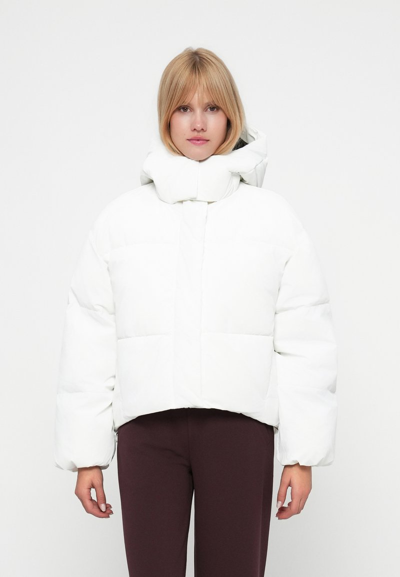 Jachetă scurtă albă tip puffer cu guler înalt, având un design matlasat, mâneci suficient de umflate și o textură netedă, lucioasă.