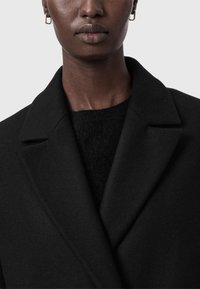 Abrigo de lana negro con cuello de solapa, que presenta una superficie suave y texturizada. Incluye un suéter negro visible debajo. Diseño simple y moderno.