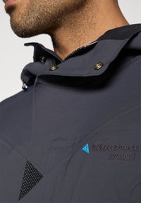 Tamsiai mėlynas atsparus vandeniui švarkas su gobtuvu, auksinėmis snap mygtukais, geometriniais raštais ir išsiuvinėtu logotipu su mėlynu akcentu trikampio pavidalu krūtinėje.