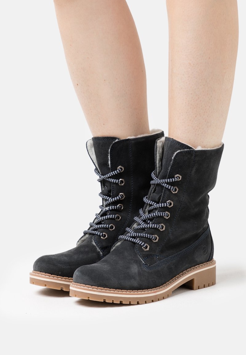 Bottes cheville en daim noir à lacets avec semelles en caoutchouc marron clair portées sur des jambes nues sur un fond blanc.