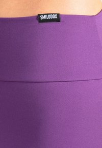 Paarse atletische tailleband gemaakt van rekbaar materiaal, met een zwart merklipje. De stof heeft een gladde textuur met een lichte glans.