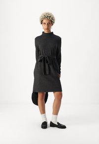 Σκούρο γκρι πλεκτό φόρεμα με ψηλό λαιμό και μακριά μανίκια, διαθέτοντας ζωνάκι στη μέση και ίσια γραμμή. Περιλαμβάνει μια υφασμάτινη μαύρη τσάντα.